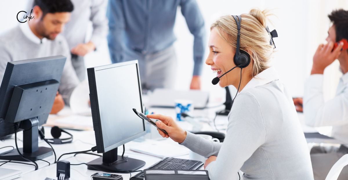 A woman talking on a headset and pointing at a computer monitor.