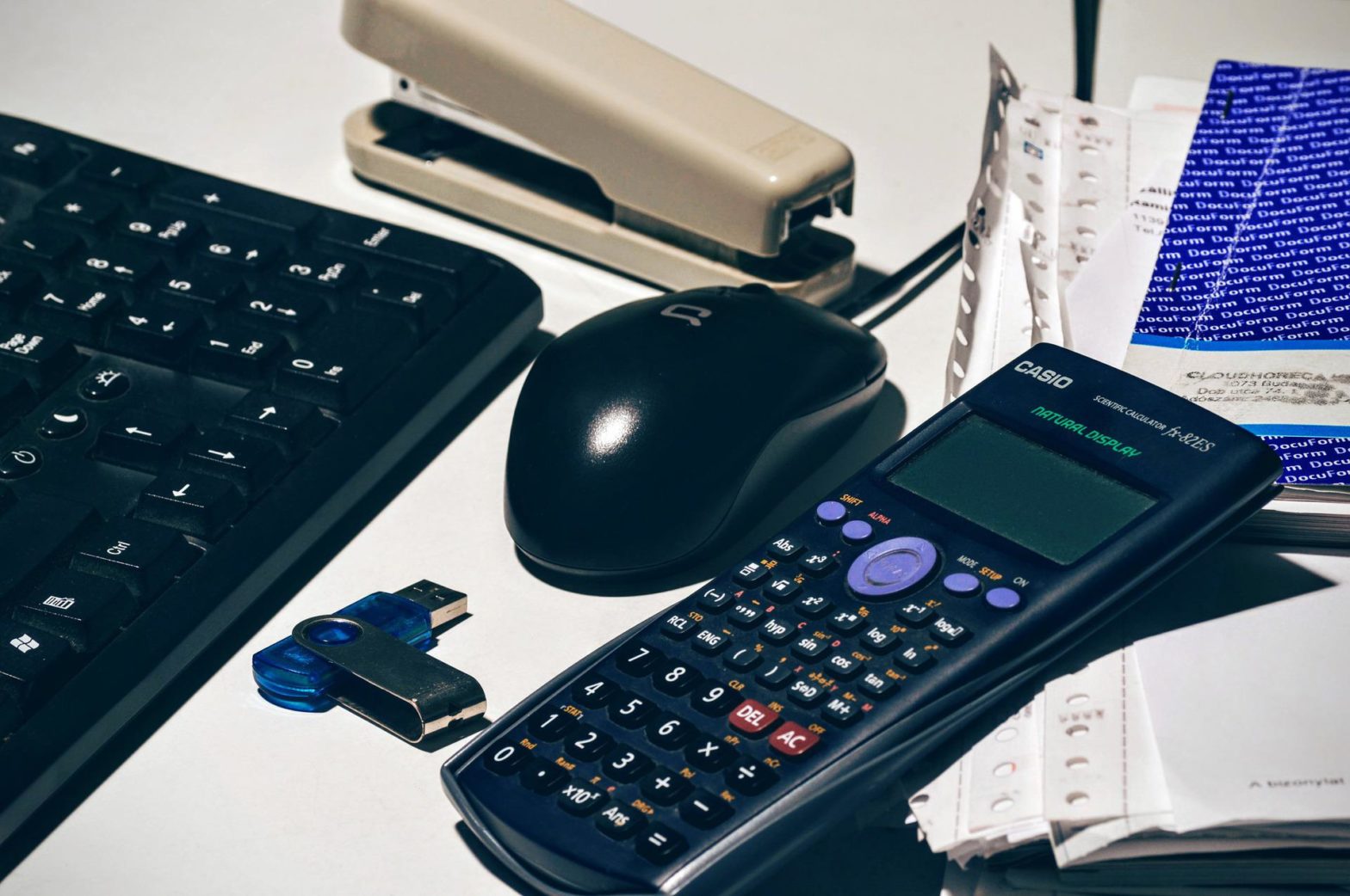 A calculator next to a computer mouse and keyboard, and stapler.