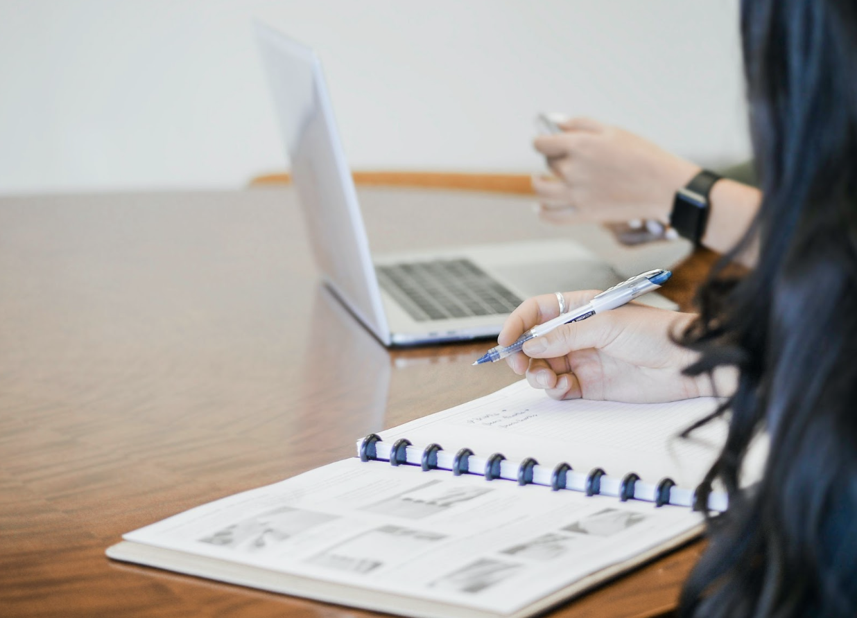 Two people writing in a notebook and working on a laptop talking about bookkeeping tasks