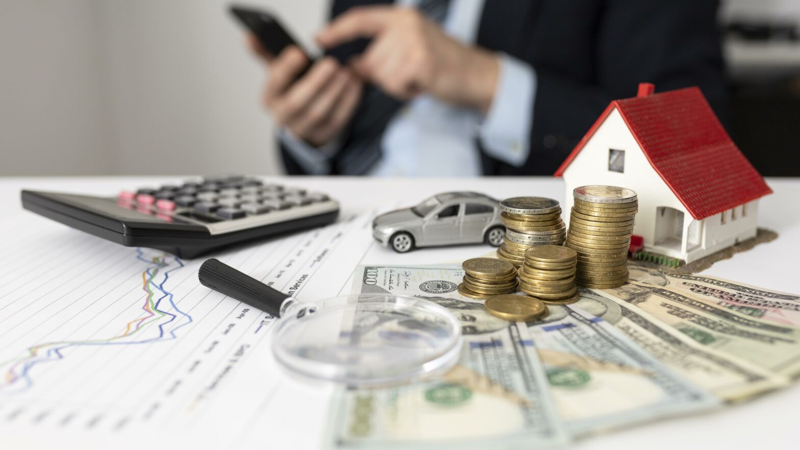A man making notes at a table with money and a calculator on it alongside a model car and house.