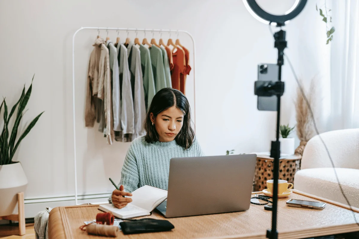 A person in a cozy workspace, writing in a notebook, with a laptop open and a ring light set up next to a clothes rack and plants.