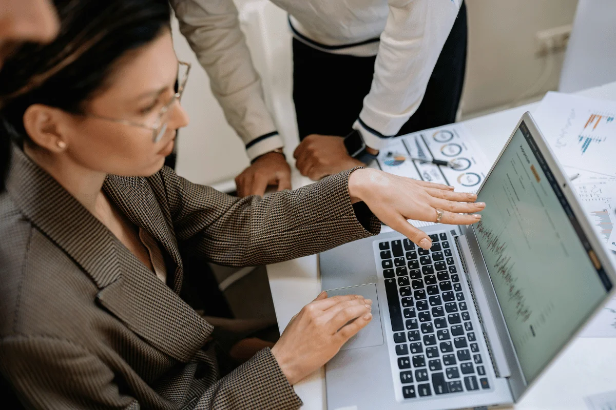 Woman pointing at financial graphs on a laptop screen with colleagues observing.
