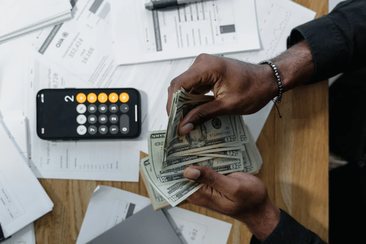 An individual counting cash with a calculator and financial papers on a wooden desk, focusing on money management.