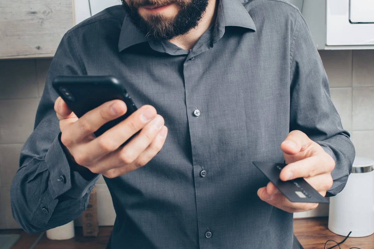 A man holds a credit card in one hand and a cell phone in the other, appearing to make a transaction. 