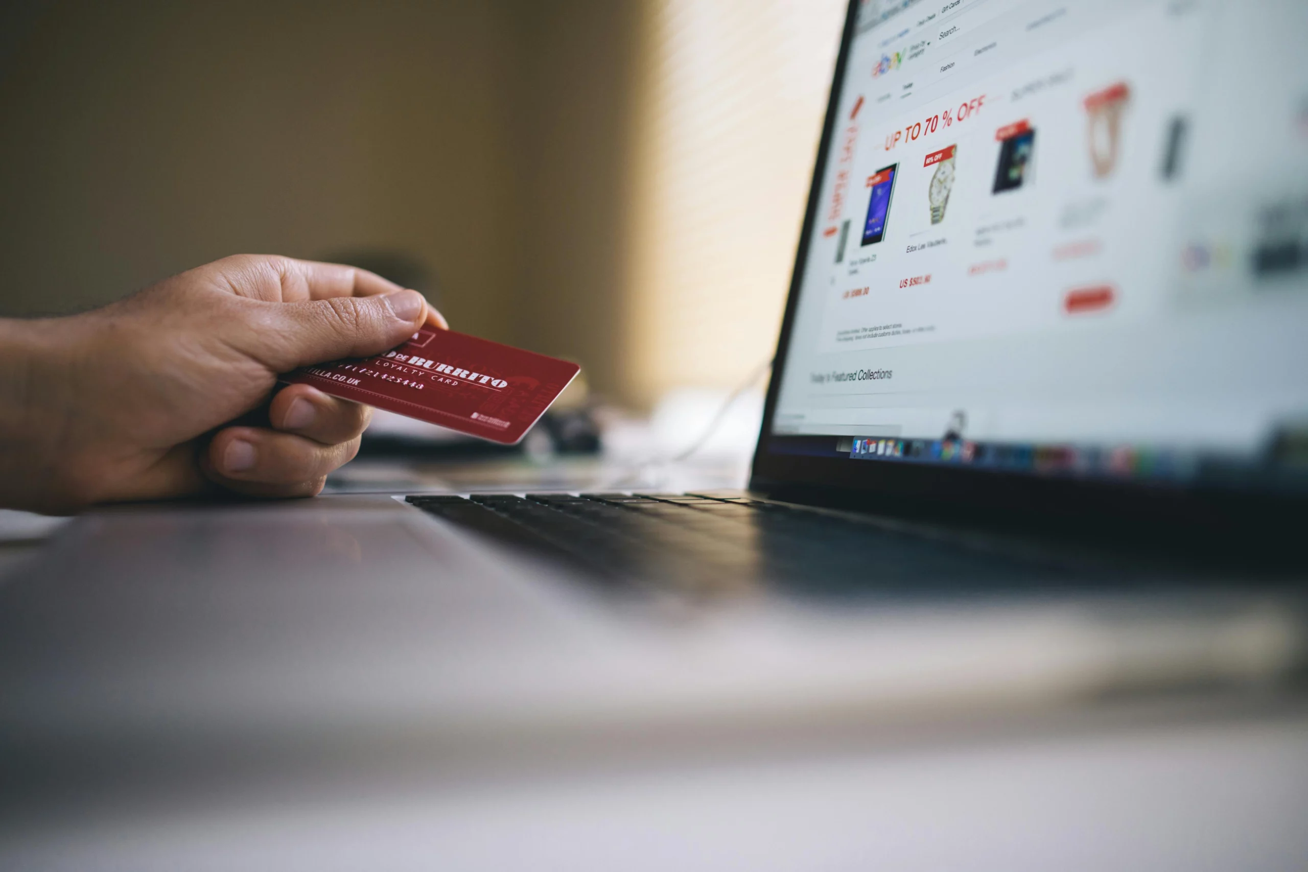 A person's hand holding a credit card in front of a laptop displaying an online shopping site.